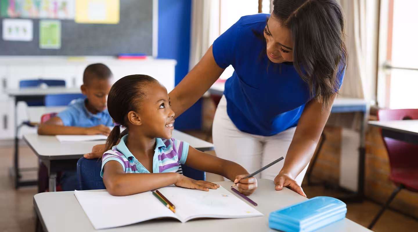 Teacher helping a young student with schoolwork in a classroom while another student works in the background.